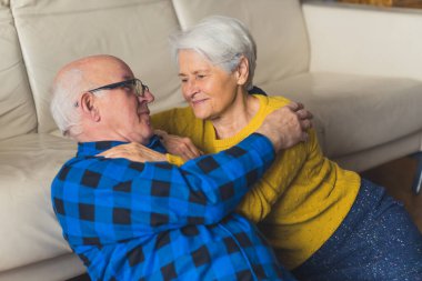 Relaxed romantic beautiful elderly family couple looking at each other and smiling while sitting on the floor near the couch in the living room at home. High quality photo