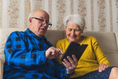 Caucasian senior man sitting with his old wife on a sofa in the living room and showing her photos on his smartphone. High quality photo