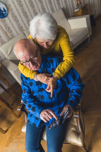 Caucasian senior confused couple trying to learn how to use a smartphone. A man sitting on a chair while a woman hugging him from behind and pointing at the screen. High quality photo