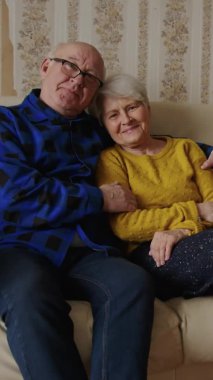 Relaxed, married elderly man and woman hugging while sitting on the couch and looking back on old times, remembering their youth. Vertical. High quality 4k footage