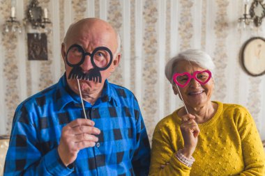 Cute joyful senior European couple holding party accessories or masks on wooden sticks - fake black mustache and pink heart-shaped glasses, laughing and having fun in the living room, enjoying