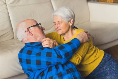 Relaxed beautiful loving embracing senior family couple looking at each other and smiling while sitting on the floor near the couch in the living room at home. High quality photo