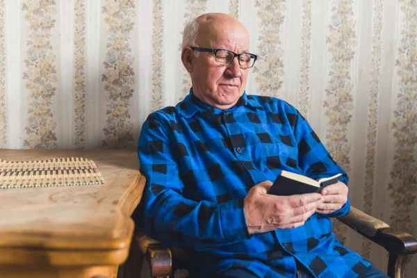 Calm European senior man sitting on a chair by the table, slowly reading a book. Life after retirement. High quality photo