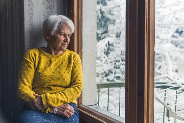 Calm caucasian senior woman sitting alone on the windowsill and looking out the window at the snowy trees, thinking about retirement. High quality photo