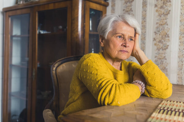 Lonely pretty senior woman, lost in thoughts, sits at the living room table with her head leaning on her arm. High-quality photo