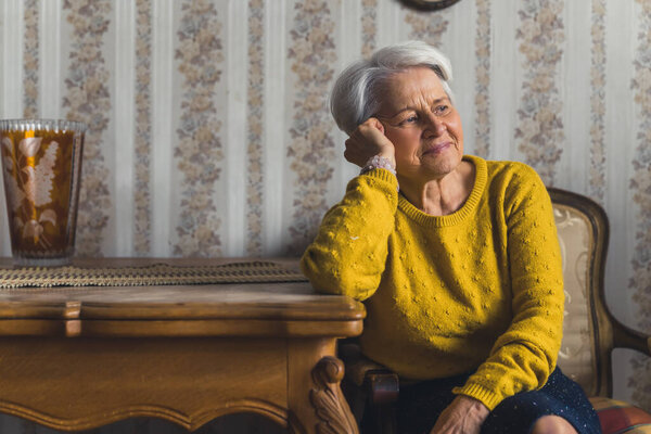 Pretty small short-haired elderly woman in a pleasant mood sitting on a chair at the living room table with her head leaning on her arm and looking away thinking. High quality photo