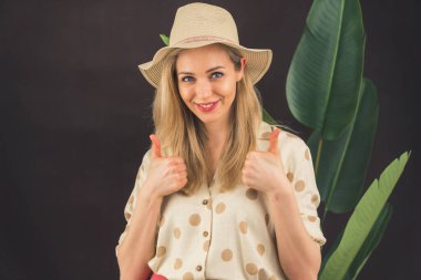 Blonde white woman wearing sun hat and summery polka-dot shirt standing next to tall plant smiling at camera showing thumbs up. Dark background studio shot. High quality photo