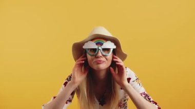 Caucasian blonde woman wearing blouse and rainbow-shaped sunglasses fixing hair and sun hat looking into camera. Studio shot. High quality 4k footage