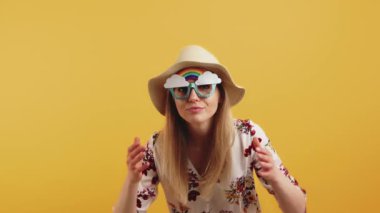 Young blonde woman wearing sun hat and rainbow glasses checking herself out in camera flipping her hair and smiling. Studio shot. High quality 4k footage