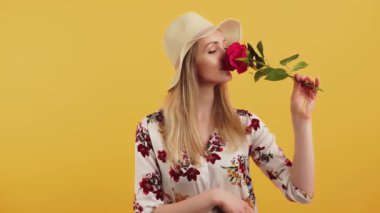 White woman with long blonde hair wearing floral blouse and sun hat acting flirty playing with rose smelling it and laughing. Studio shot. High quality 4k footage