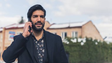 Hispanic man with slick hair and beard wearing shirt and coat walking around town talking on the phone. Business call. High quality photo