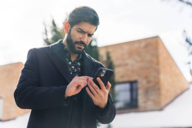 Hispanic bearded businessman wearing coat outside on winter morning swiping through messages on smarphone. High quality photo