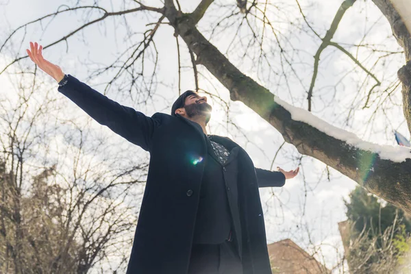 Bearded man wearing suit and coat outstretched arms celebrating outside around trees looking at blue sky and shining sun . High quality photo