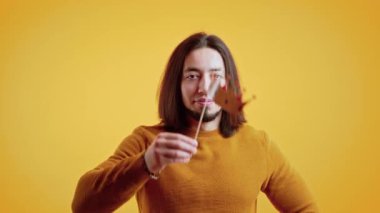 Caucasian man with long brown hair wearing orange sweater holding paper crown above head and sticking tongue out looking into camera. Yellow background. High quality 4k footage