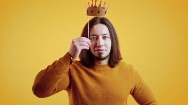 Young white brunette man with beard wearing orange sweater putting fake paper crown above head smiling looking into camera. Yellow background. High quality 4k footage
