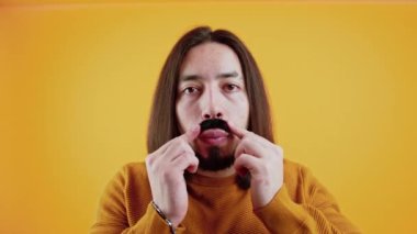 Caucasian dark-haired man with beard looking focused into camera glueing on fake black mustache. Yellow background. High quality 4k footage