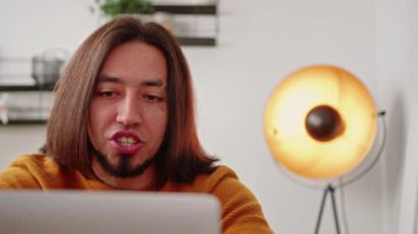 White dark-haired man with beard wearing orange sweater participating in online meeting speaking and looking at laptop. White room indoor shot. High quality 4k footage