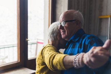 Small senior woman dancing with her husband in the living room and resting her head on his shoulder, holding hands, spending time together during cold winter days. High quality photo