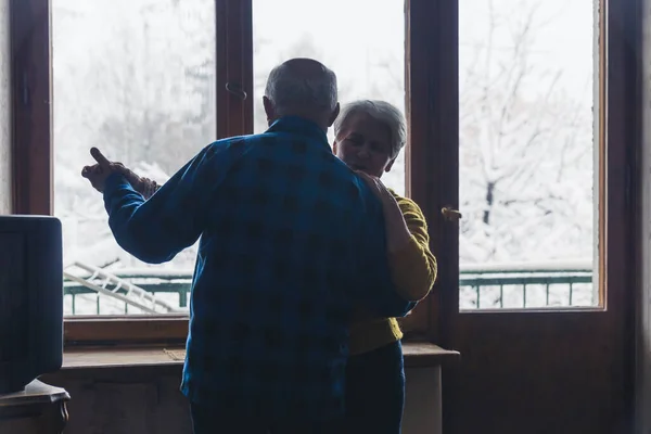 Sweet senior European married couple hugging and dancing in the dark living room, holding hands, enjoying retirement together during cold winter days. High quality photo