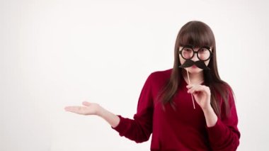 Caucasian brunette woman with bangs wearing red sweater holding paper glasses with mustache disguise in front of her face. Horizontal video. Isolated copy space. High quality 4k footage