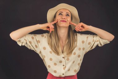 attractive stylish positive blonde lady with a hat looking up and touching her cheeks with her fingers - black background. High quality photo