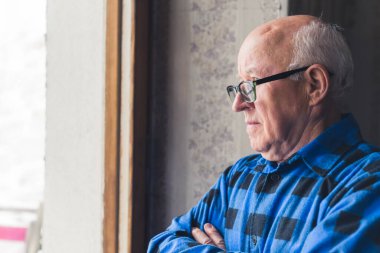Retired senior bald man with arms crossed, wearing his glasses and thoughtfully looking out the window. High quality photo