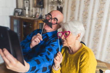 Cute joyful senior European couple holding party accessories or masks on wooden sticks - fake black mustache and pink heart-shaped glasses, laughing and taking selfies on a phone, enjoying retirement