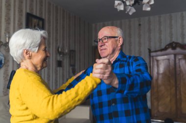 Happy romantic old married couple smiling and dancing in the living room, holding hands, having fun at home. High quality photo