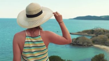 Vacation in Greece. A slim caucasian tanned woman in a striped dress and a beach hat standing on a rock cliff near the crystal clear water of the endless sea. High quality 4k footage