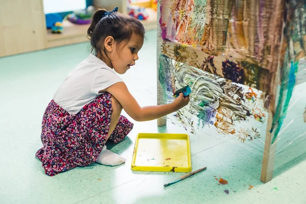 Cling film painting. Little girl toddler painting with a sponge and paints on a cling film wrapped all the way round the wooden shelf unit. Creative activity for kids development at the nursery school