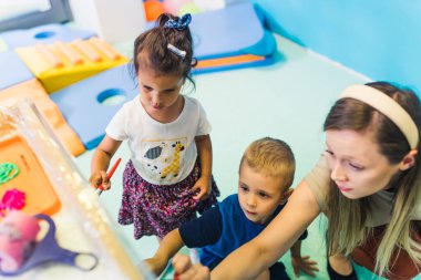 Cling film painting. Toddler painting with a sponge, brushes and paints on a cling film wrapped all the way round the wooden shelf unit. A teacher helping them. Creative activity for kids sensory