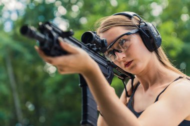 Young caucasian woman wearing protective goggles and headphones aiming submachine gun on training ground. Blurred background horizontal shot. High quality photo