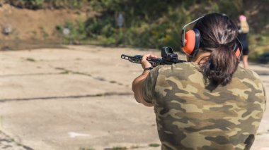 Rear view of man with ponytail wearing camo t-shirt and safety headphones practicing aim using submachine gun. Outdoor shooting range. Horizontal shot. High quality photo