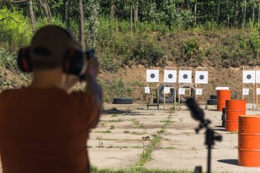 Rear view of man in safety headphones next to field glass practicing with hand gun on firing range. Target practice. Focus on background. Outdoor horizontal shot. High quality photo
