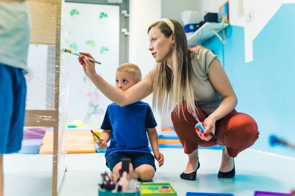 Young caucasian day-care-center teacher showing toddlers how to paint objects with a brush and paints on a cling film wrapped around the wooden shelving. Fun activity for kids imagination and brain