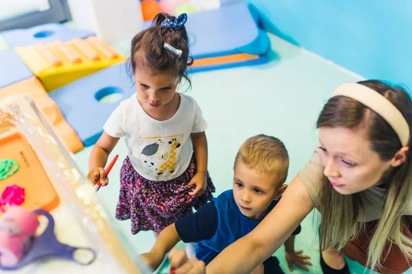 Cling film painting. Toddler painting with a sponge, brushes and paints on a cling film wrapped all the way round the wooden shelf unit. A teacher helping them. Creative activity for kids sensory
