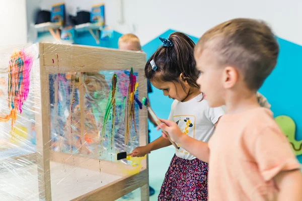 Cling film painting. Toddler painting with a sponge, brushes and paints on a cling film wrapped all the way round the wooden shelf unit. A teacher helping them. Creative activity for kids sensory