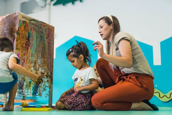 Cling film painting. Toddler painting with a sponge, brushes and paints on a cling film wrapped all the way round the wooden shelf unit. A teacher helping them. Creative activity for kids sensory