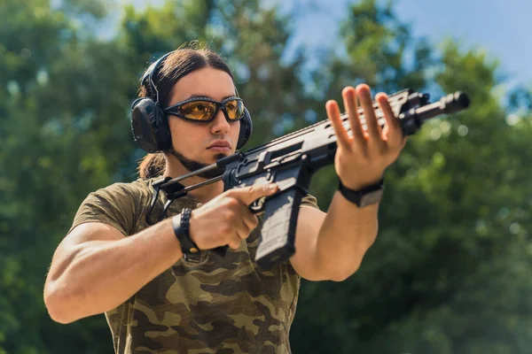 Bearded man in safety goggles headphones and camo t-shirt practicing using submachine gun. Outdoor firing range. Horizontal shot. High quality photo