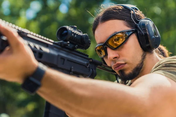 White man with beard in protective goggles and headphones aiming submachine gun. Outdoor firing range. Horizontal shot. High quality photo