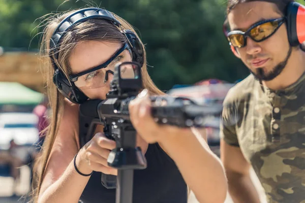 Caucasian woman in safety headphones and goggles aiming submachine gun watched by bearded man in camouflage t-shirt. Firearms training ar fiiring range. Horizontal shot. High quality photo