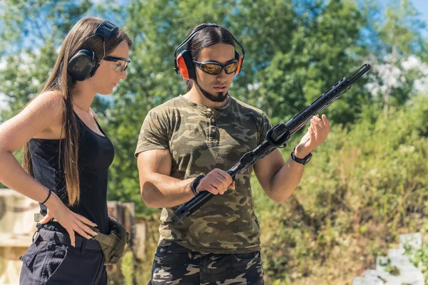 Woman observing man in camo t-shirt using submachine gun. Firearms training outdoor shooting range. Client with instructor in safety headphones and goggles. Horizontal shot. High quality photo