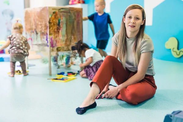 Young caucasian day-care-center teacher sitting on the floor and looking at the camera while on the background toddlers painting with tempera paints on a cling film wrapped around the wooden shelving