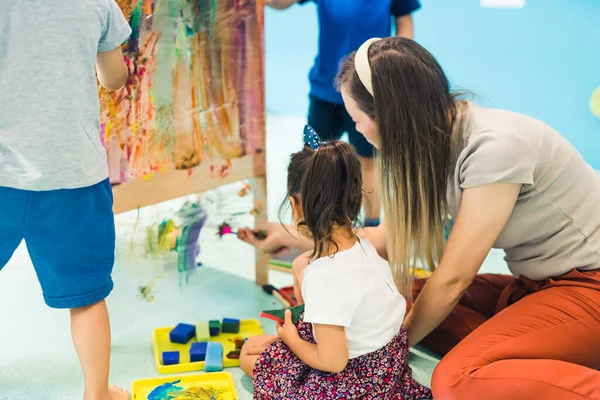 Cling film painting. Toddler painting with a sponge, brushes and paints on a cling film wrapped all the way round the wooden shelf unit. A teacher helping them. Creative activity for kids sensory