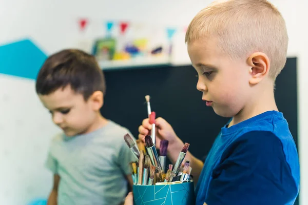 Nursery school. Toddlers having art game class, engaged in the process, blonde toddler boy holding a jar with paint brushes. Fun art activity for kids fine and gross skills, creativity and imagination