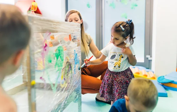 Cling film painting for improving kids imagination. Toddlers painting with, brushes, rollers and paints on a cling film wrapped around the wooden shelving stand. Fun activity for kids sensory skills