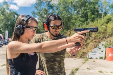 Male instructor showing female client how to aim handgun. Safety goggles and headphones. Firearms training at outdoor shooting range. Horizontal shot. High quality photo
