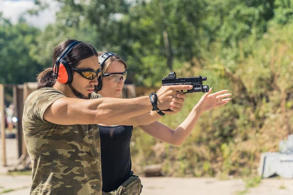 White man in camo t-shirt safety headphones and goggles holding handgun with female instructor showing how to aim it. Firearm training ar firing range. Outdoor horizontal shot. High quality photo