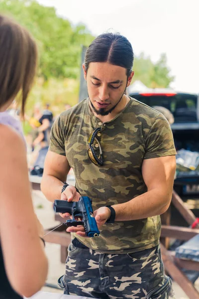 Caucasian muscular man wearing camo t-shirt presenting a handgun to female client. Buying and selling firearms topic. Outdoor vertical shot. High quality photo