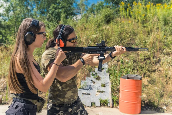 Woman standing next to man aiming submachine gun both wearing safety headphones and goggles. Firearms training at firing range. Outdoor horizontal shot. High quality photo